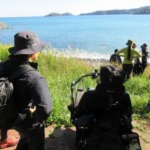 7名が浜辺から海を眺めている後ろ姿。青い海の奥には山や小さな島が見える。