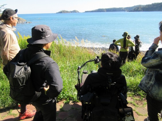 7名が浜辺から海を眺めている後ろ姿。青い海の奥には山や小さな島が見える。